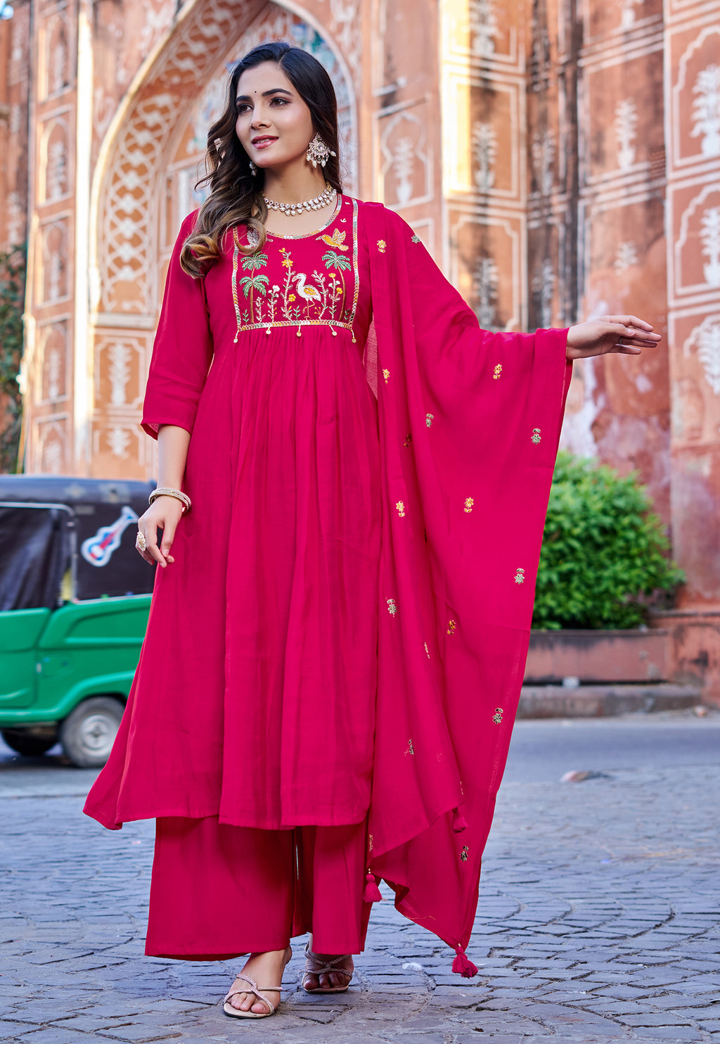 Woman in a bright pink traditional outfit with embroidery standing in front of an architectural background.
