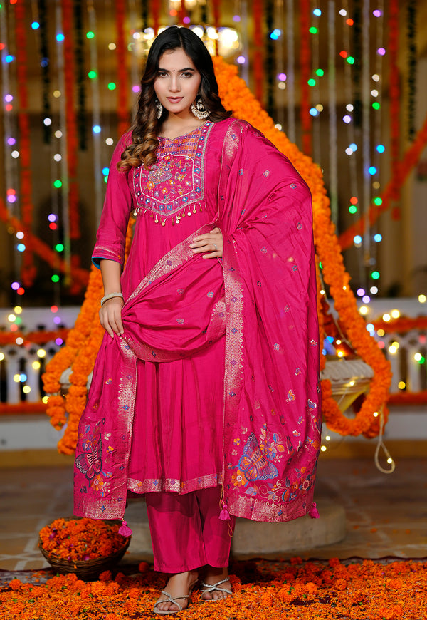 A woman modeling a bright pink traditional Indian outfit with matching dupatta, decorated with floral embroidery and mirror work, set against a festive background with marigold garlands and multicolored string lights.