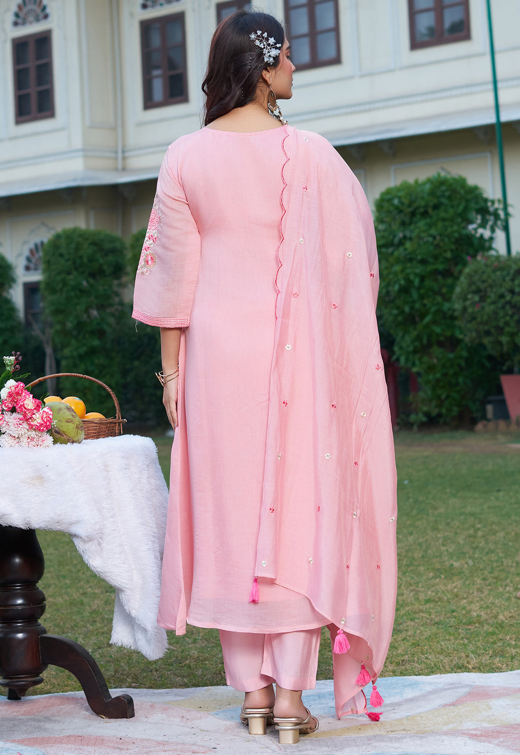 Woman in a pink traditional outfit standing outdoors with a table and flowers in the background.