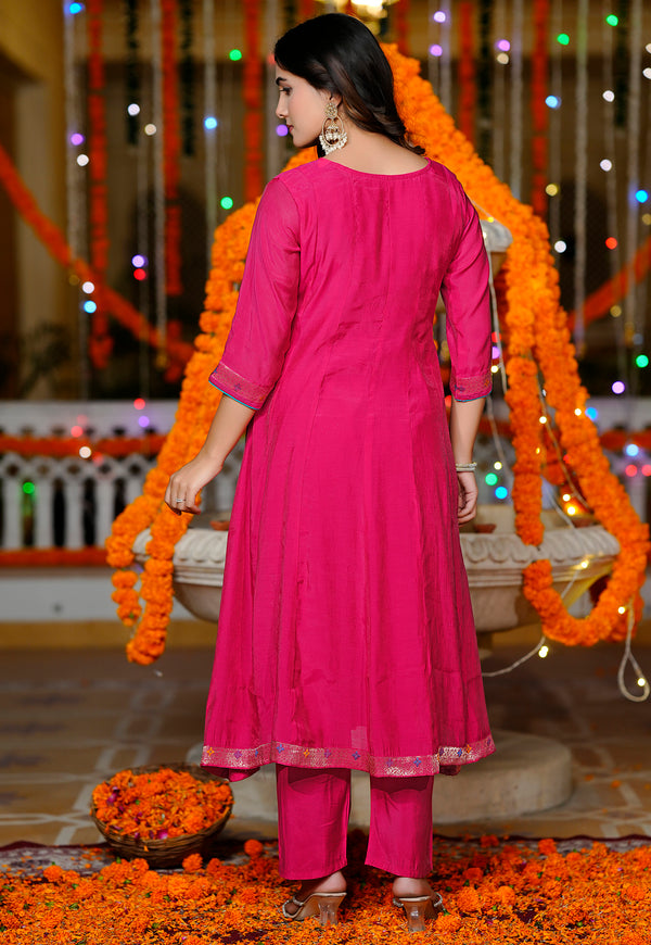 A woman wearing a bright pink traditional Indian outfit with three-quarter sleeves and matching pants, standing in a decorated indoor setting with orange marigold garlands and string lights.