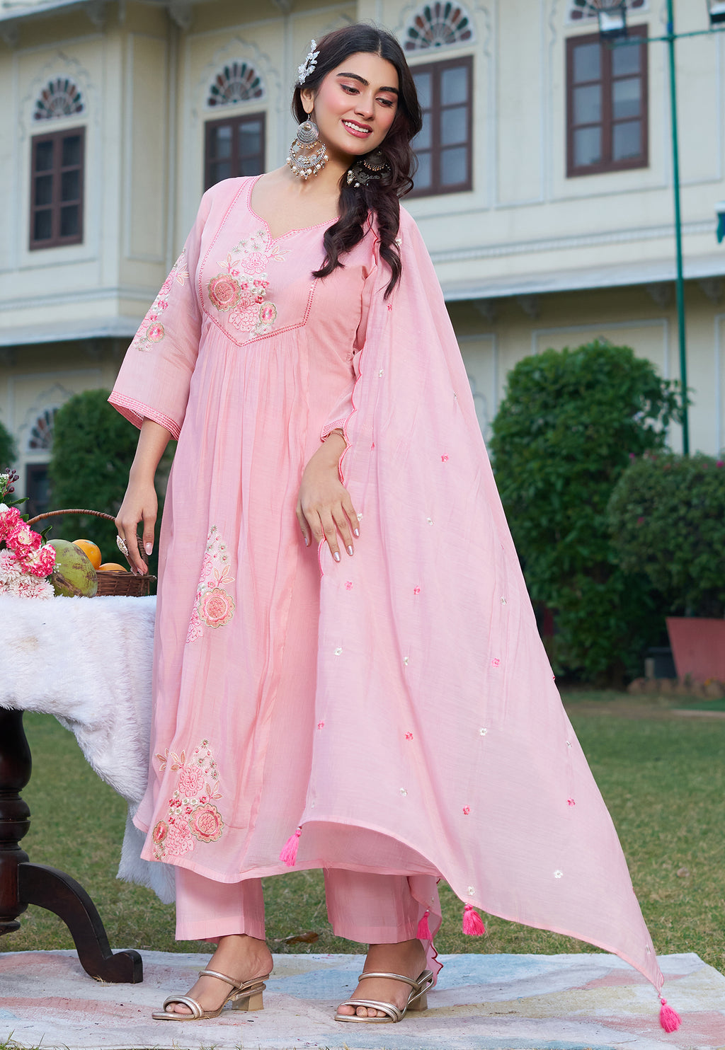 Woman in a pink traditional outfit standing outdoors with a building and greenery in the background.