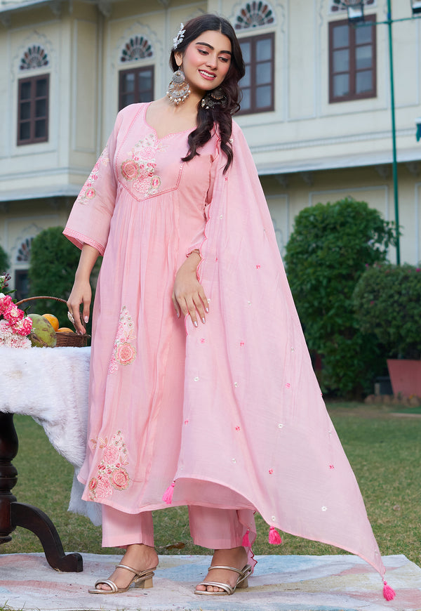 Woman in a pink traditional outfit standing outdoors with a building and greenery in the background.