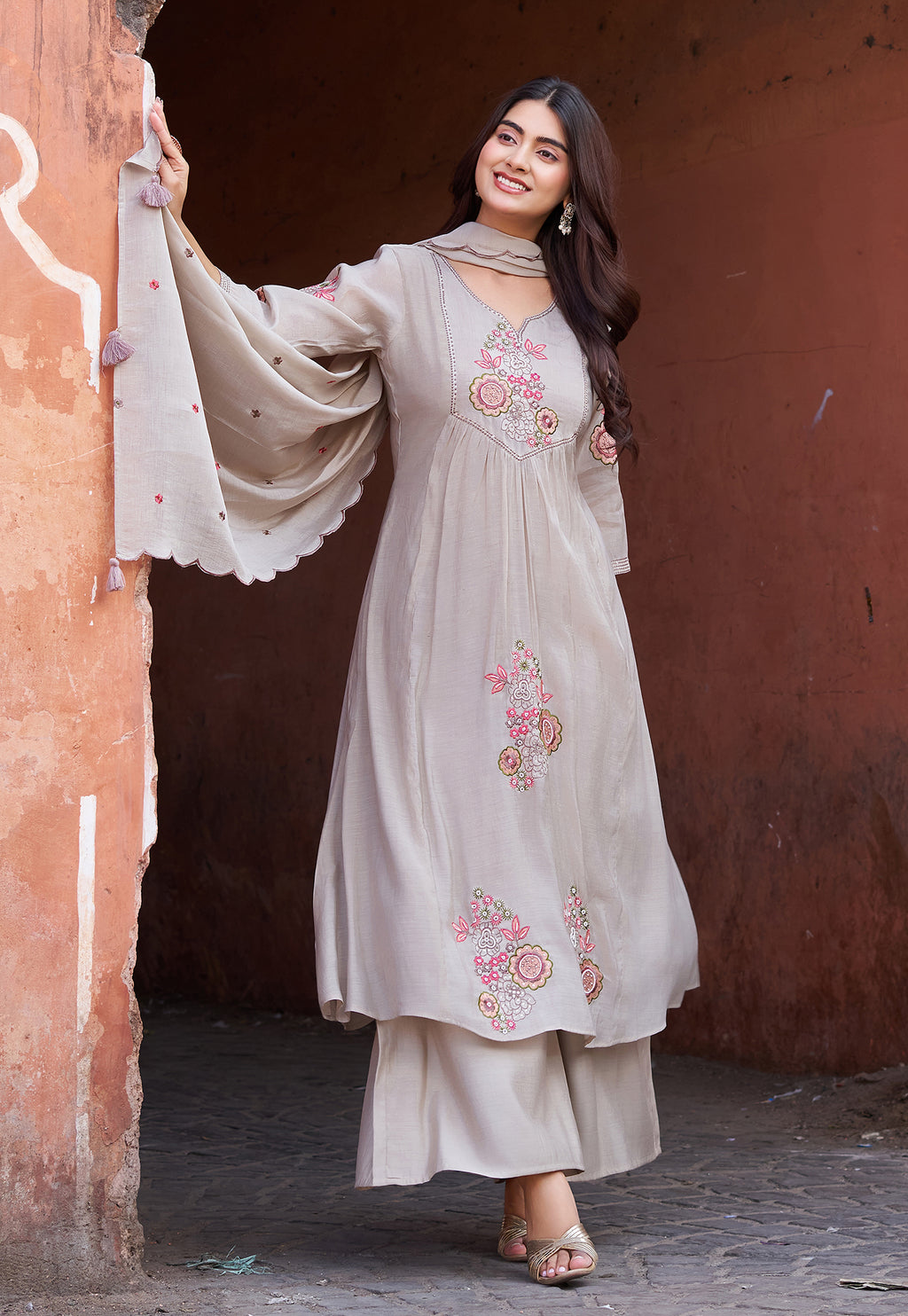Woman in a light-colored embroidered outfit standing in front of a reddish-brown wall.