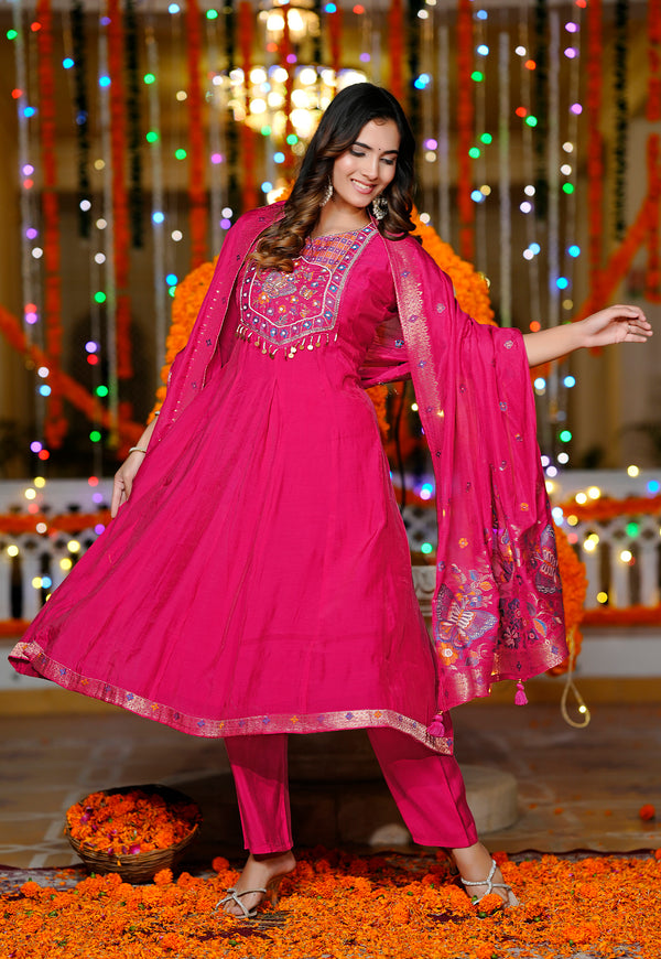 A woman modeling a bright pink traditional Indian salwar kameez suit with matching dupatta, decorated with intricate embroidery on the chest, standing on a floor covered with orange marigold petals in front of a festive backdrop with hanging lights and garlands.