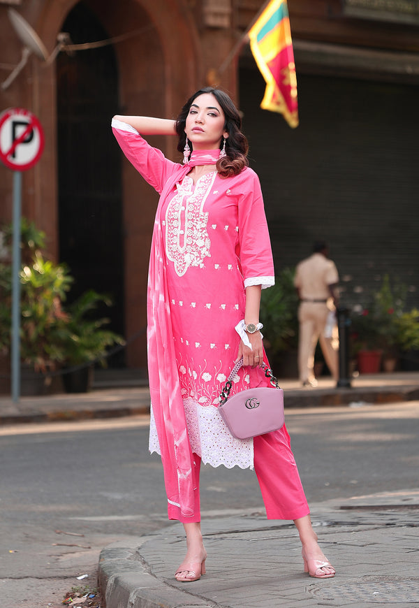 A woman stands on a city street wearing a bright pink traditional outfit with white embroidery and white lace trim, holding a pink handbag.