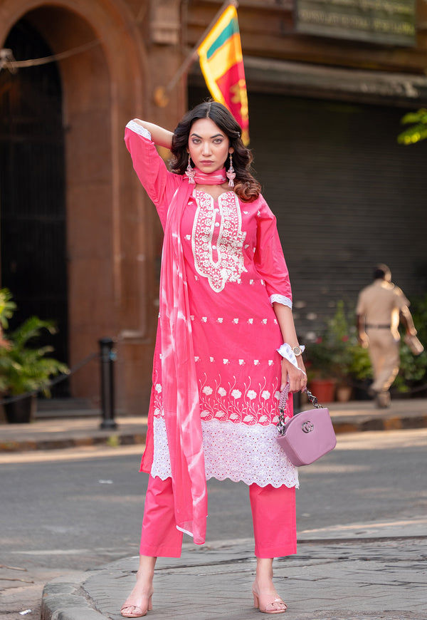 A woman modeling a bright pink traditional Indian salwar kameez set with white lace embroidery, holding a matching pink handbag in an outdoor street setting.