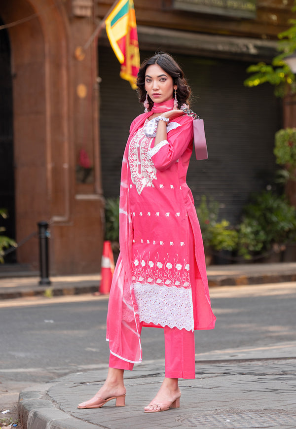 A woman models a bright pink three-piece traditional outfit with white embroidery and a matching dupatta in an outdoor urban setting.