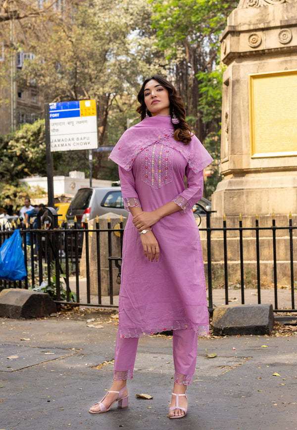 A woman modeling a lavender purple kurta set with matching pants and pinkish-beige sandals, standing outdoors in front of a stone monument and trees.