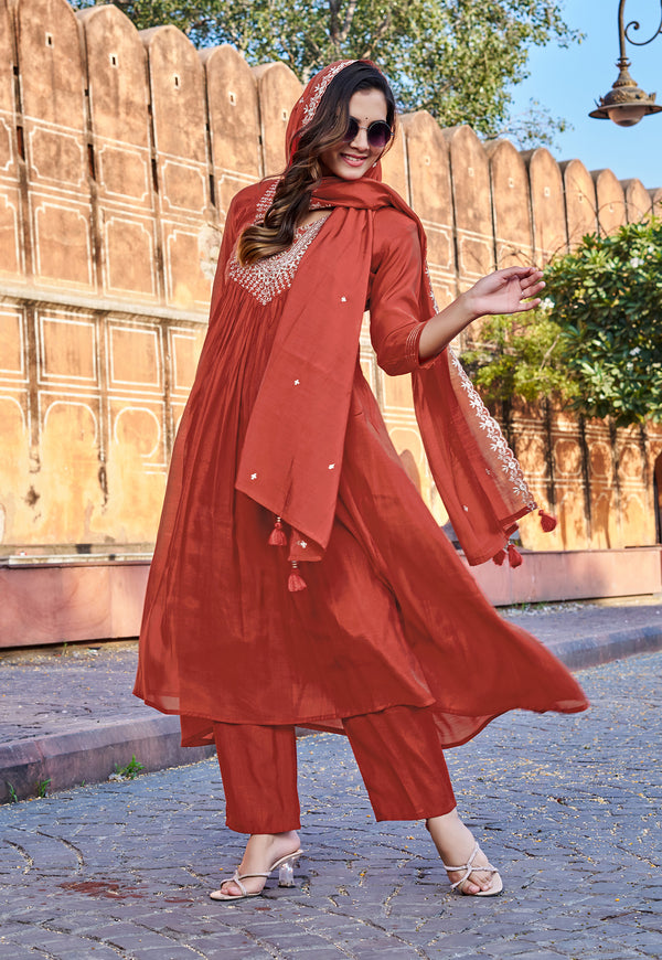 A woman modeling a rust-orange three-piece salwar kameez set with a matching dupatta, featuring silver embroidery on the chest and sleeve, set against a background of a wooden fort wall and cobblestone street.