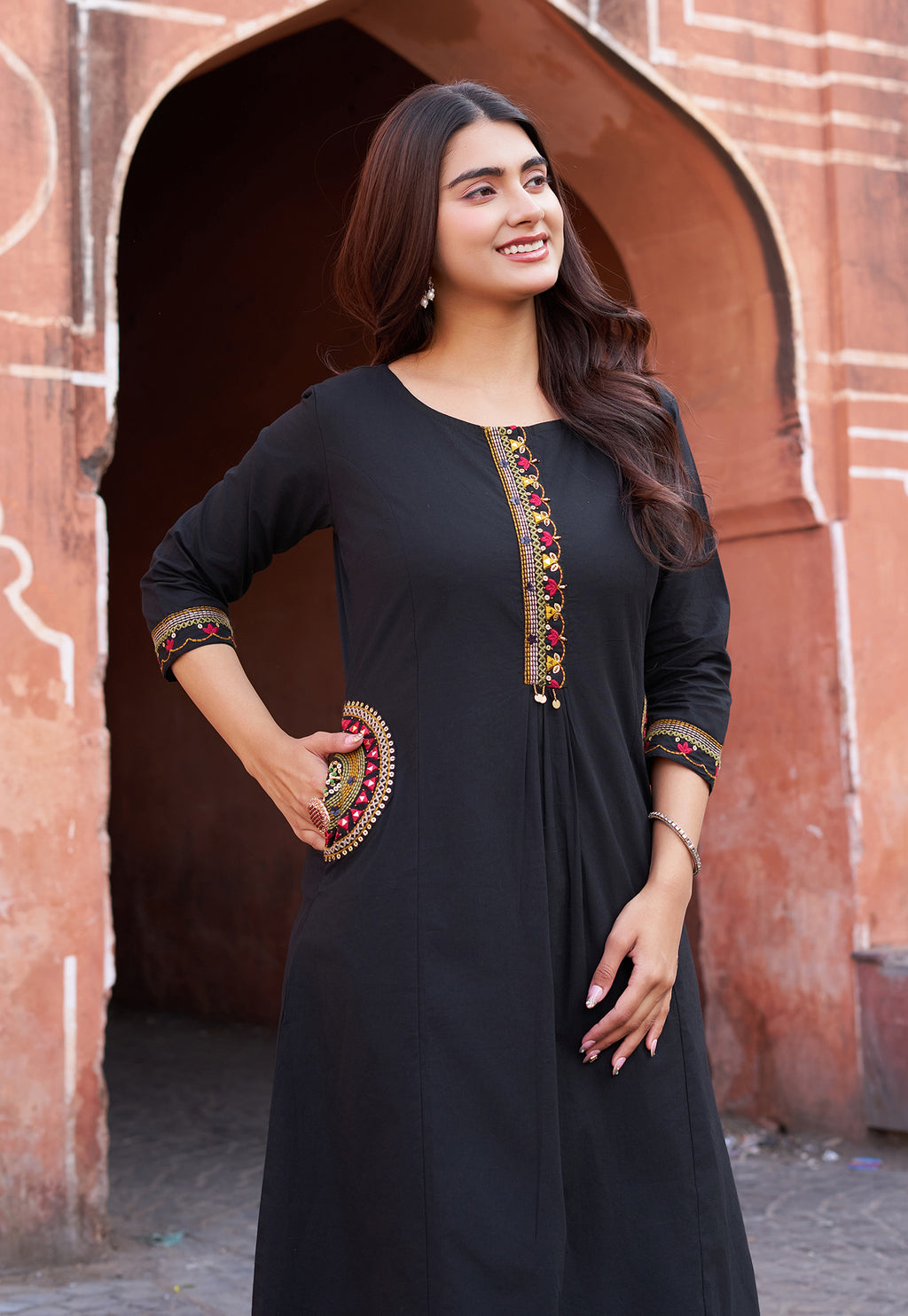 Woman wearing a black dress with colorful embroidery standing in front of an archway.