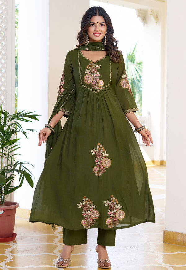 Woman wearing a green embroidered outfit in a bright room with plants.
