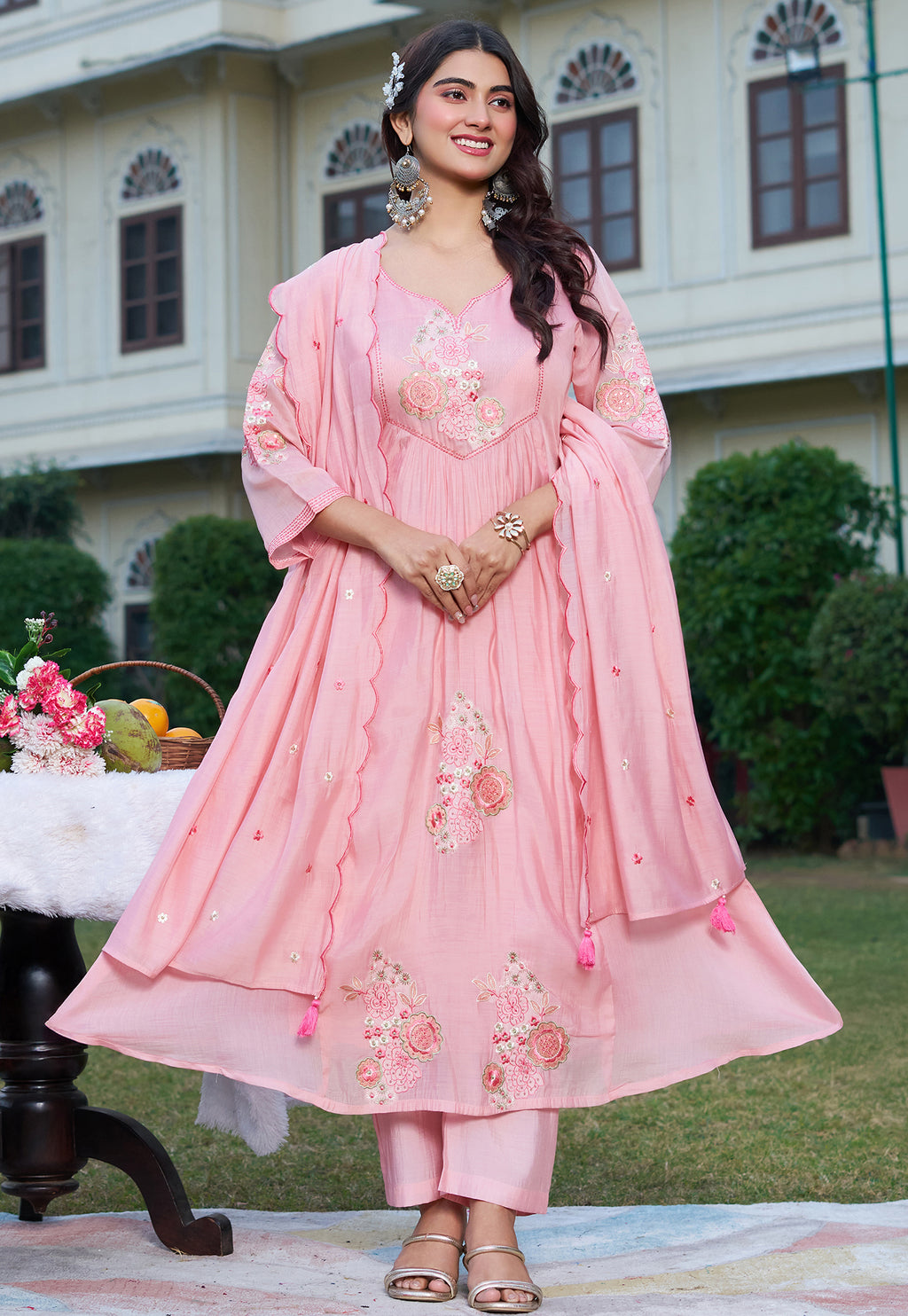 Woman in a pink traditional outfit standing outdoors with a building and greenery in the background