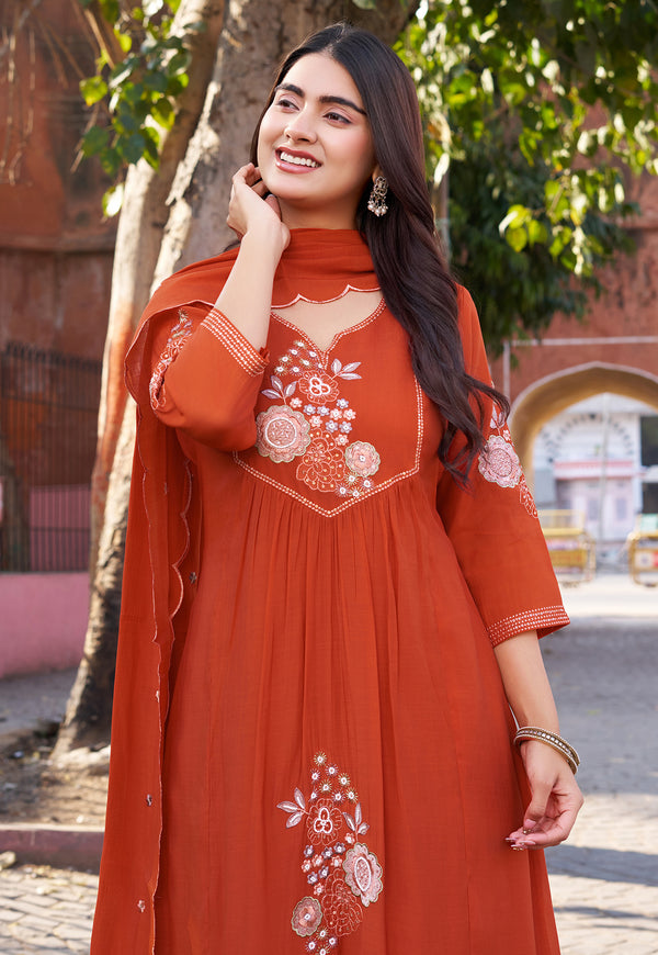 Woman wearing an orange embroidered dress standing outdoors with trees and a building in the background.
