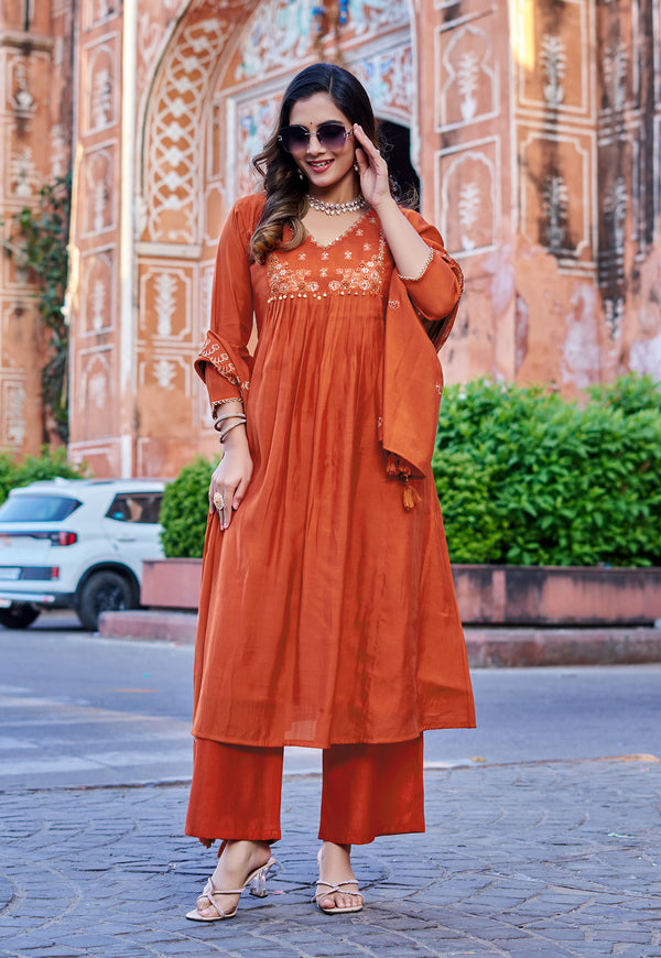 A woman models a rust-orange kurta set with matching wide-leg pants and a draped dupatta in front of a historic pink sandstone building.