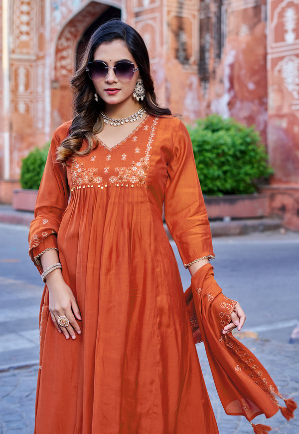 A woman wearing a rust-orange long kurta with embroidered neckline and matching dupatta, standing outdoors in front of a historic stone building.