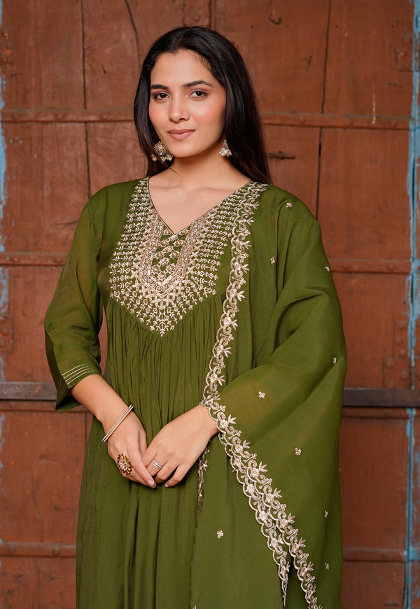 A woman wearing an olive green traditional outfit with intricate gold embroidery, standing in front of a rustic brown wooden door.