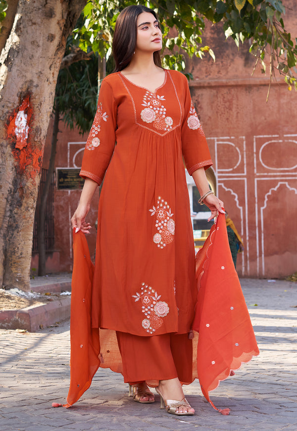 Woman wearing an orange embroidered traditional outfit standing outdoors with a tree and architectural background.