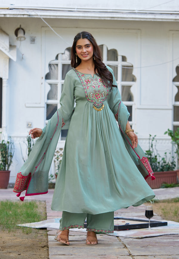 A woman modeling a mint green traditional Indian salwar kameez suit with floral embroidery on the chest and dupatta, standing outdoors in front of a white house.