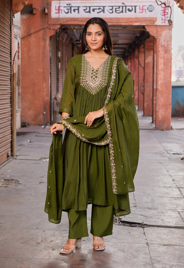 A woman models an olive green three-piece salwar kameez suit with gold embroidery, standing in a rustic outdoor alleyway.