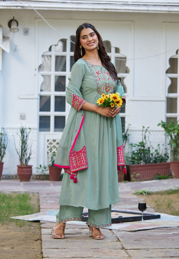 A woman stands outdoors in front of a white building, wearing a sage green traditional kurta set with pink and gold embroidery, holding a bouquet of yellow sunflowers and wearing a matching pink embroidered shoulder bag.