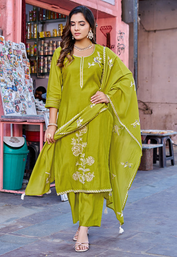 A woman models a lime green ethnic three-piece suit with white floral embroidery, featuring a kurta, matching pants, and a dupatta, set against a street market background.