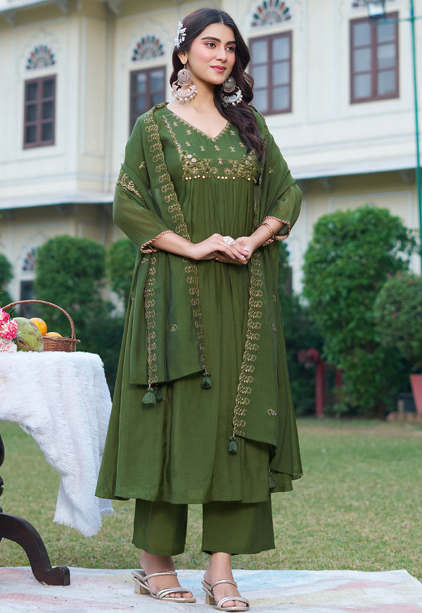 A woman models an olive green traditional Indian salwar kameez suit with gold embroidery, standing outdoors in front of a white building and green trees.