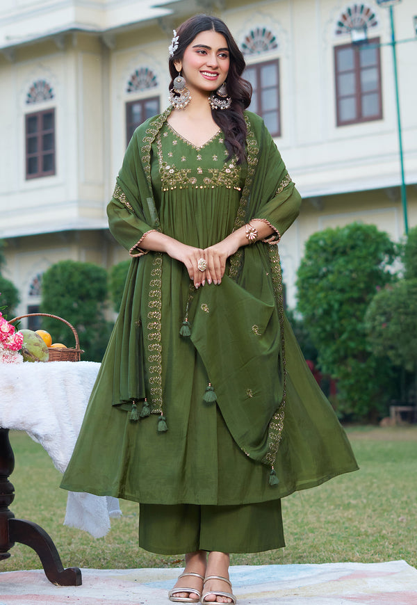 A woman models an olive green ethnic salwar kameez suit with gold embroidery in an outdoor garden setting. The set includes a kurta, matching trousers, and a draped dupatta.
