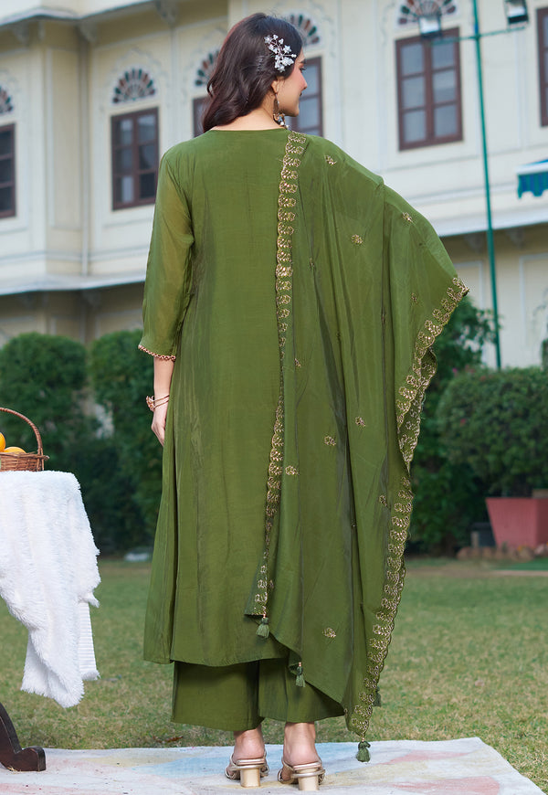 A woman stands outdoors facing away from the camera wearing an olive green three-piece traditional outfit with a matching dupatta featuring gold embroidery.